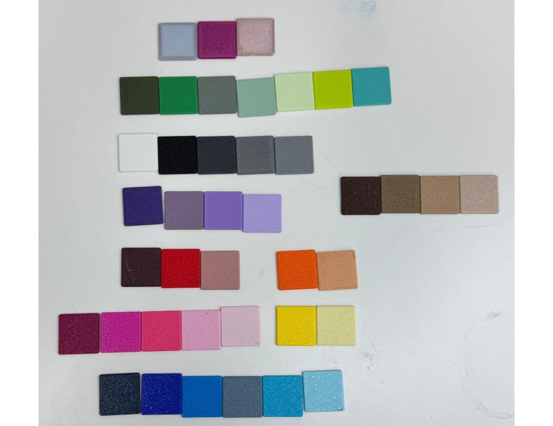 Op de afbeelding: Een verzameling kleine, vierkante tegels gerangschikt in rijen, met een grote verscheidenheid aan effen kleuren, waaronder tinten blauw, groen, rood, geel, paars, roze, oranje, bruin, zwart, wit en grijs met een matte of licht getextureerde afwerking.