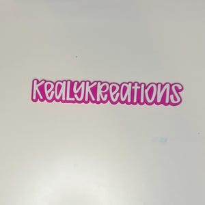 May include: A pink and white sticker with the text "KealyKreations" in a playful, handwritten font. The sticker has a white outline, and the text is in a bright pink color. The sticker is likely used for branding or decoration.