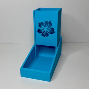 May include: A bright blue dice tower and tray set. The tower features a cut-out flower design. The tray is rectangular and connected to the base of the tower. The set is made of a solid, smooth material.