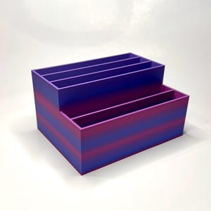 May include: A two-tiered organizer in shades of purple and pink. The rectangular box has multiple compartments, ideal for storing office supplies or craft materials. The color gradient creates a visually appealing design.