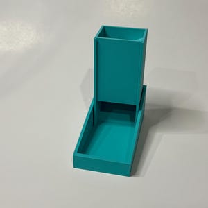 May include: A teal-colored dice tower and tray set. The tower is rectangular with an open top, and the tray is a matching teal color. The set is made of a solid material and is designed for tabletop gaming.