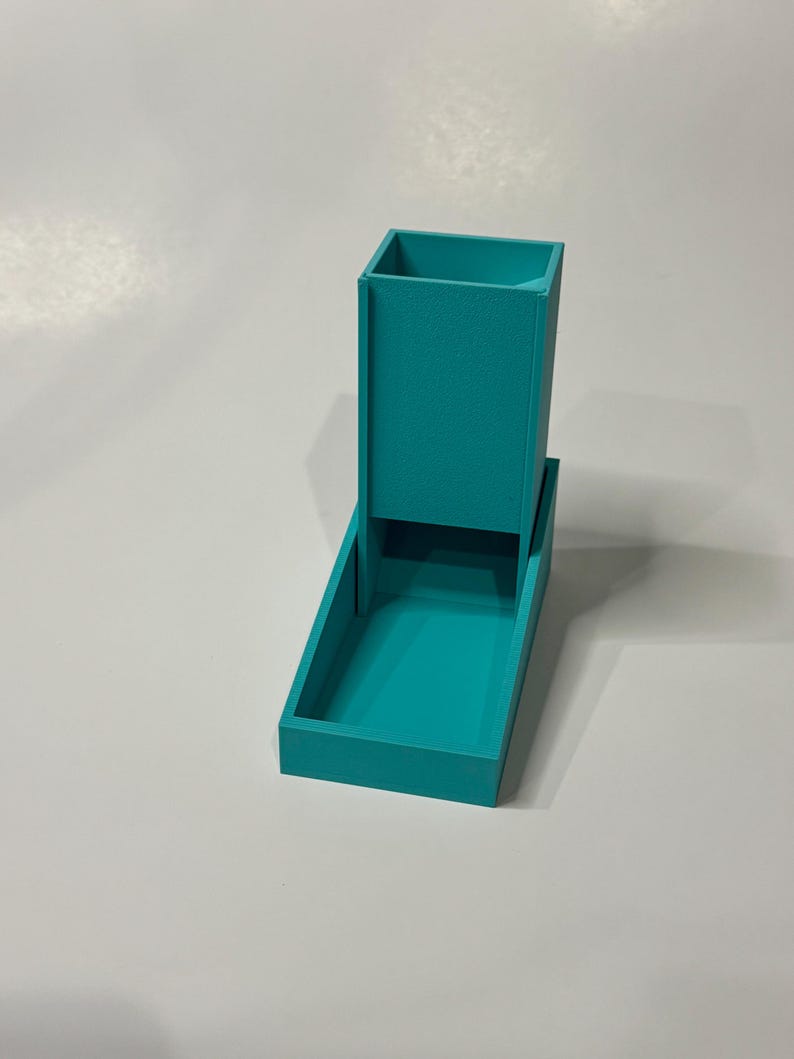 Op de afbeelding: Een turquoise dobbelsteentoren met een hoge verticale schacht en een bevestigd rechthoekig dienblad aan de basis. De structuur heeft een matte afwerking en een eenvoudig, functioneel ontwerp bedoeld voor het rollen van dobbelstenen.