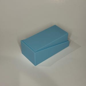 Op de afbeelding: Een rechthoekige lichtblauwe doos met een bijpassend deksel dat in een hoek is geplaatst. De doos heeft een gladde, matte afwerking en een strak, geometrisch ontwerp. Het deksel rust enigszins scheef op de basis, waardoor de binnenrand zichtbaar is.