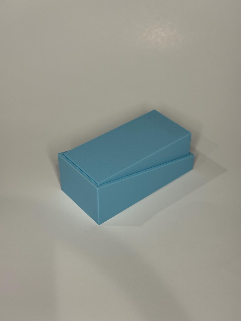 Op de afbeelding: Een rechthoekige lichtblauwe doos met een bijpassend deksel dat in een hoek is geplaatst. De doos heeft een gladde, matte afwerking en een strak, geometrisch ontwerp. Het deksel rust enigszins scheef op de basis, waardoor de binnenrand zichtbaar is.