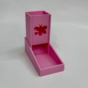 May include: A bright pink dice tower featuring a cutout of a butterfly on the front panel. The structure consists of a tall vertical rectangular chamber for dropping dice and an attached lower tray for catching them, all in a solid matte finish.