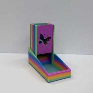 Op de afbeelding: Een regenboogkleurige dobbelsteentoren met een verticale rechthoekige schacht met een uitgesneden vlindervorm op het voorpaneel. De structuur bevat een aan de basis bevestigd bakje voor het opvangen van dobbelstenen, afgewerkt in kleurverloop.