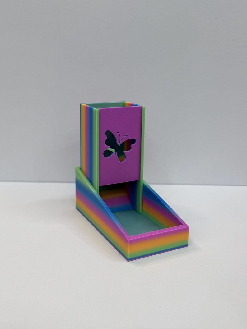 Op de afbeelding: Een regenboogkleurige dobbelsteentoren met een verticale rechthoekige schacht met een uitgesneden vlindervorm op het voorpaneel. De structuur bevat een aan de basis bevestigd bakje voor het opvangen van dobbelstenen, afgewerkt in kleurverloop.