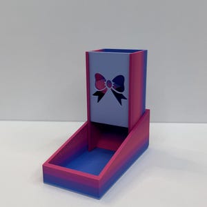 Op de afbeelding: Een dobbelsteentoren met een verticale rechthoekige schacht en een voorste bakje, uitgevoerd in een kleurverloop van roze, paars en blauw. Het voorpaneel van de toren toont een uitgesneden strikontwerp dat in het midden is geplaatst.