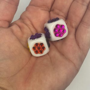 May include: Two white cubic dice featuring embedded circular clusters of colorful rhinestones on each visible face. One die displays a bright orange rhinestone flower pattern, while the other shows a vibrant pink rhinestone flower design.