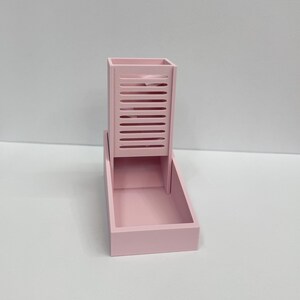 Op de afbeelding: Een lichtroze dobbelsteentoren met een verticale rechthoekige schacht met horizontale sleuven aan de voorzijde en een vierkante basis. De structuur is gemaakt van een stevig materiaal met een matte afwerking en een consistente pastelroze kleur.