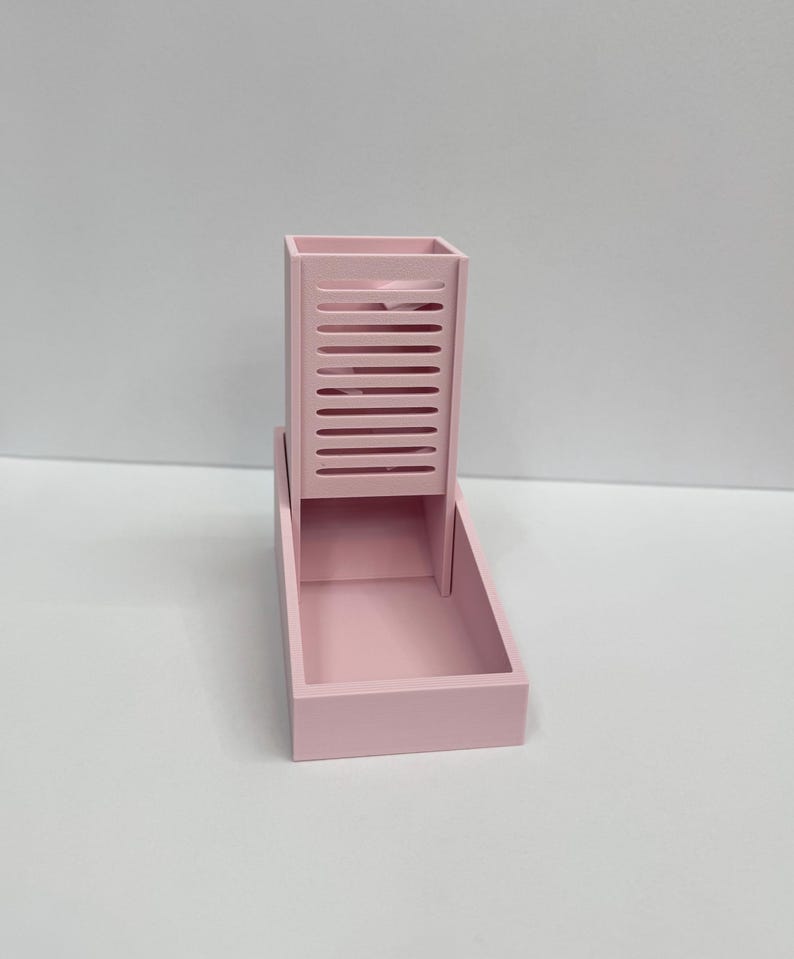 Op de afbeelding: Een lichtroze dobbelsteentoren met een verticale rechthoekige schacht met horizontale sleuven aan de voorzijde en een vierkante basis. De structuur is gemaakt van een stevig materiaal met een matte afwerking en een consistente pastelroze kleur.