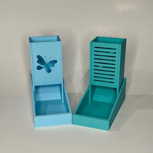 May include: Two wooden desk organizers in shades of blue and teal. The blue organizer has a butterfly cutout, while the teal one features horizontal slats. Both have a rectangular base and a tall, open compartment.