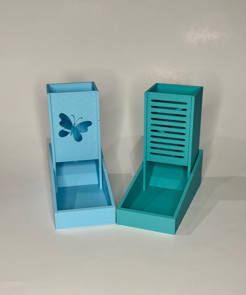 Op de afbeelding: Twee dobbelsteentorens met open bakjes aan de basis. De linker toren is lichtblauw met een vlinderuitsnede aan de voorzijde. De rechter toren is talingblauw met horizontale latten aan de voorzijde. Beide zijn rechtopstaande, rechthoekige structuren.