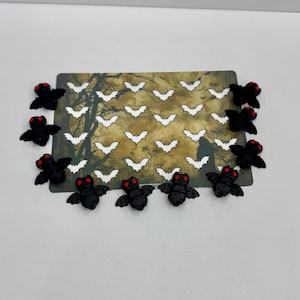 May include: A Halloween-themed card with a forest scene and white bat silhouettes. Small, black bat embellishments with red eyes surround the card, creating a festive and eerie atmosphere. Ideal for Halloween decorations or crafts.
