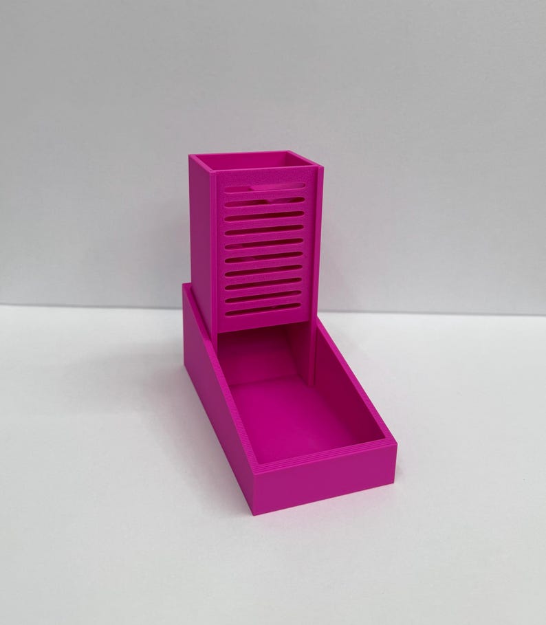 Op de afbeelding: Een felmagenta 3D-geprinte dobbelsteentoren met een verticale rechthoekige kolom met horizontale sleuven aan de voorzijde en een open bak aan de basis om dobbelstenen op te vangen. De structuur is solide, egaal van kleur en glad afgewerkt.
