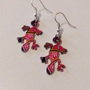 May include: A pair of silver dangle earrings featuring a pink and black gecko design. The gecko has a patterned design with white, yellow, and black accents.