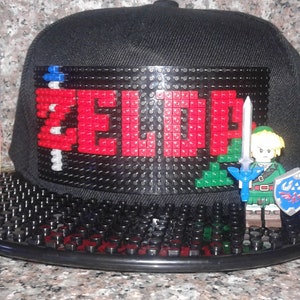 May include: Black baseball cap with a LEGO design featuring the word "ZELDA" in red and green LEGO bricks. A LEGO figure of Link from the Legend of Zelda video game series is attached to the side of the cap.