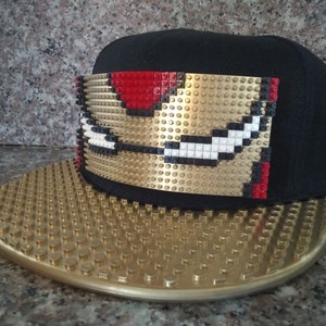 May include: A black and gold baseball cap with a gold Lego design on the front. The cap has a gold Lego brim.