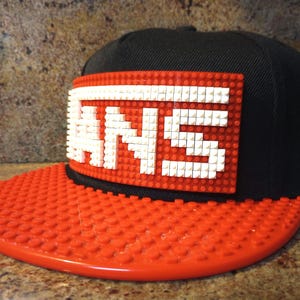 May include: A black and red baseball cap with a white Lego brick design that spells out "VANS" on the front.