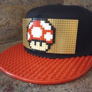 May include: A black and gold baseball cap with a red and white pixelated mushroom design on the front. The brim of the cap is red and made of plastic.