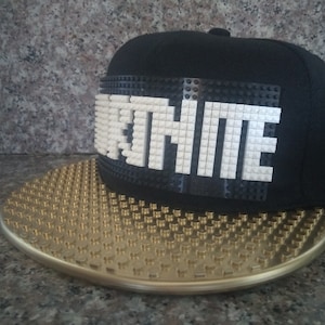 May include: A black and gold baseball cap with a white LEGO brick design that spells out "FORTNITE".