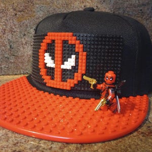 May include: A black and red baseball cap with a Deadpool logo made of red and black Lego bricks. A small Deadpool Lego minifigure is attached to the front of the cap.