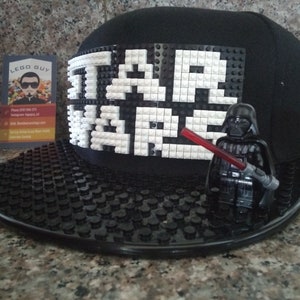 Op de afbeelding: Zwarte baseballcap met een platte klep, met de woorden "STAR WARS" in witte Lego-stenen. Een Lego Darth Vader-figuur met een rood lichtzwaard staat op de klep. De cap ligt op een getextureerd oppervlak.