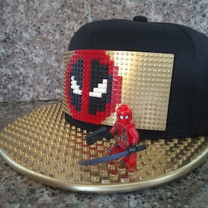 May include: A black and gold baseball cap with a Deadpool logo made of Lego bricks. A small Deadpool Lego minifigure is standing on the cap's brim.