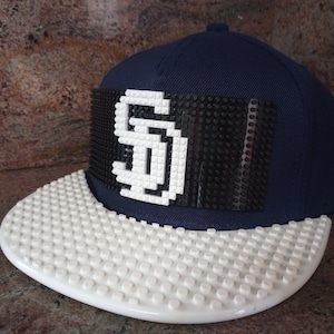 May include: Navy blue baseball cap with a white and black design. The front of the cap features a white "SD" logo on a black background. The brim is white and textured. A stylish accessory for sports fans.