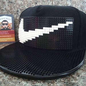 May include: Black baseball cap with a white Nike swoosh logo constructed from black and white Lego bricks. The brim of the cap is also covered in Lego studs. A business card for "Lego Guy" is in the background.