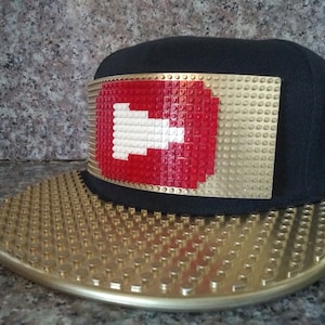 May include: A black and gold baseball cap with a red and white Lego-style play button on the front.