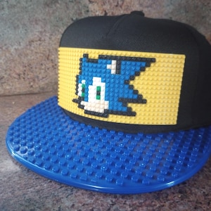 May include: A black and blue baseball cap with a yellow and blue Lego-style design of a video game character. The cap has a blue Lego-style brim.