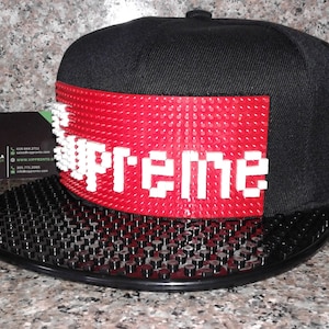 May include: Black baseball cap with a red panel featuring the white word "Supreme". The cap has a flat brim with a textured surface. The cap is displayed on a speckled surface.
