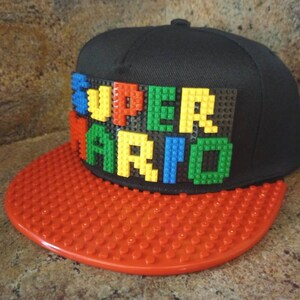 May include: A black and red baseball cap with a LEGO brick design that spells out "SUPER MARIO" in colorful bricks.