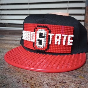 May include: A black and red baseball cap with a red and white Lego design featuring the text "STATE".