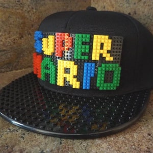 May include: Black and white baseball cap with a black brim and a black crown. The crown has a LEGO-style design that spells out "SUPER MARIO" in colorful plastic bricks.