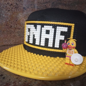 May include: A black and yellow baseball cap with a white LEGO brick design that says "NAF" on the front. A yellow toy figure with a pink dress and a cupcake is standing on the cap.