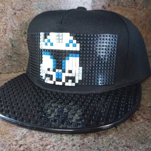 May include: Black snapback hat with a pixelated design of a white and blue character on the front. The brim and the design area are textured. The hat is a classic, flat-brim style.