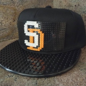 Könnte beinhalten: Schwarze Baseballkappe mit einem schwarzen und weißen Buchstaben "S" aus Lego-Steinen auf der Vorderseite. Der Schirm der Kappe ist ebenfalls aus Lego-Steinen.