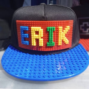 May include: A black baseball cap with a blue brim. The front of the cap features a name spelled out in colorful Lego bricks, "ERIK".