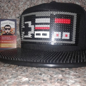 Peut inclure: Casquette de baseball noire avec un design LEGO noir et gris d'une manette de jeu vidéo sur le devant. La casquette a un dos en maille noire et une sangle réglable noire.