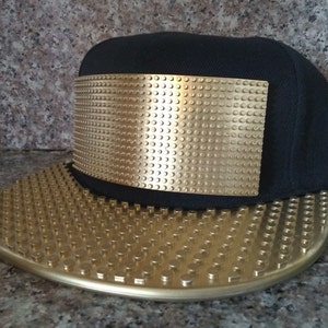 May include: A black baseball cap featuring a rectangular gold-colored panel on the front and a flat brim, both covered in a raised, grid-like pattern of small circular studs that resemble building block bricks across the entire surface.