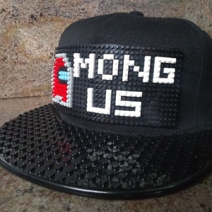 May include: A black baseball cap with a black and white Lego-style patch that says "Among Us". The bill of the cap is also covered in black Lego-style studs.