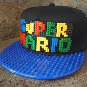 May include: A black and blue baseball cap with a LEGO brick design. The front of the cap features the words "SUPER MARIO" spelled out in colorful LEGO bricks.