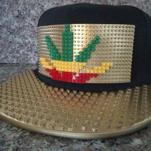 May include: A black and gold baseball cap with a green, yellow, and red marijuana leaf design made of Lego bricks.