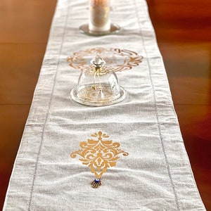 May include: A white linen table runner with gold decorative designs and a blue evil eye charm. The runner is on a wooden table with a glass cloche and a white candle.