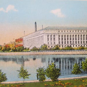 May include: Vintage postcard featuring a large, white building with numerous windows and columns, set against a blue sky with a few clouds. The building is reflected in the calm water of a river, with trees and green grass in the foreground.