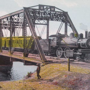 May include: Vintage image of a steam train crossing a metal bridge over water. The train is black with white wheels and a yellow passenger car. The bridge is dark gray with the number "96" on top. A person walks on the bank.