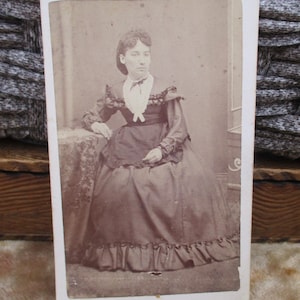 May include: A black and white photograph of a woman wearing a dark dress with a white lace collar and a ruffled skirt. She is sitting on a chair with her hands in her lap.
