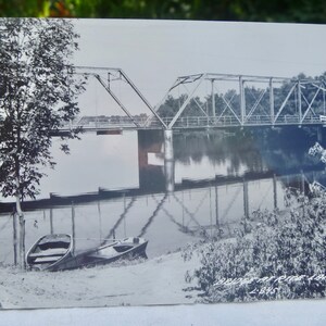 May include: Black and white vintage postcard showing a bridge over a river with two boats docked on the shore. The text on the postcard reads "Bridge at Rice Lake, Wis. - 1942".